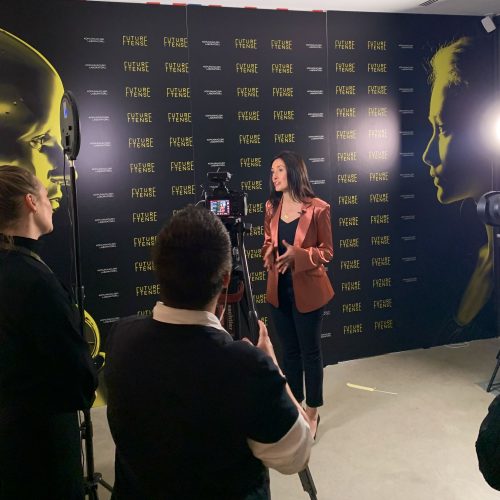 Erica Orange standing in front of a press wall speaking to cameras at the Future Tense Conference